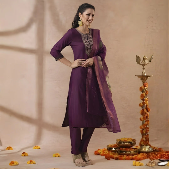 Woman in a purple traditional outfit standing next to decorative items on a beige background