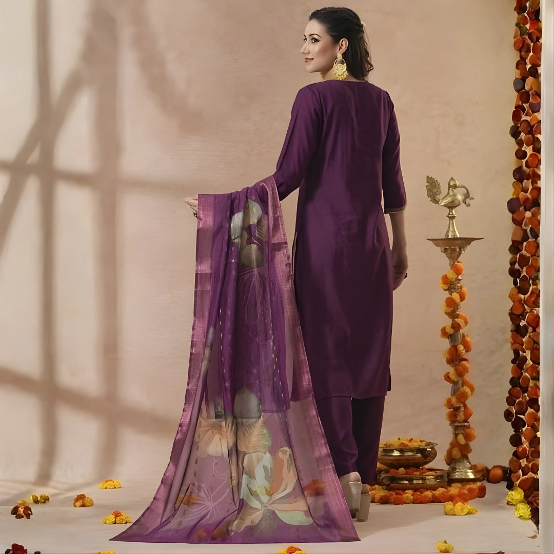 Woman in a purple traditional outfit with a decorative scarf, standing against a beige wall with decorative elements.