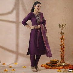 Woman in a purple traditional outfit standing next to decorative items on a beige background
