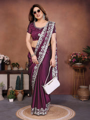 Woman in a purple saree with white floral patterns holding a white handbag, standing against a brown wall with decorative plants.