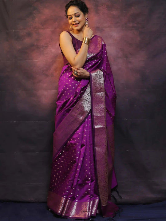 Woman wearing a purple saree with a silver border against a plain background