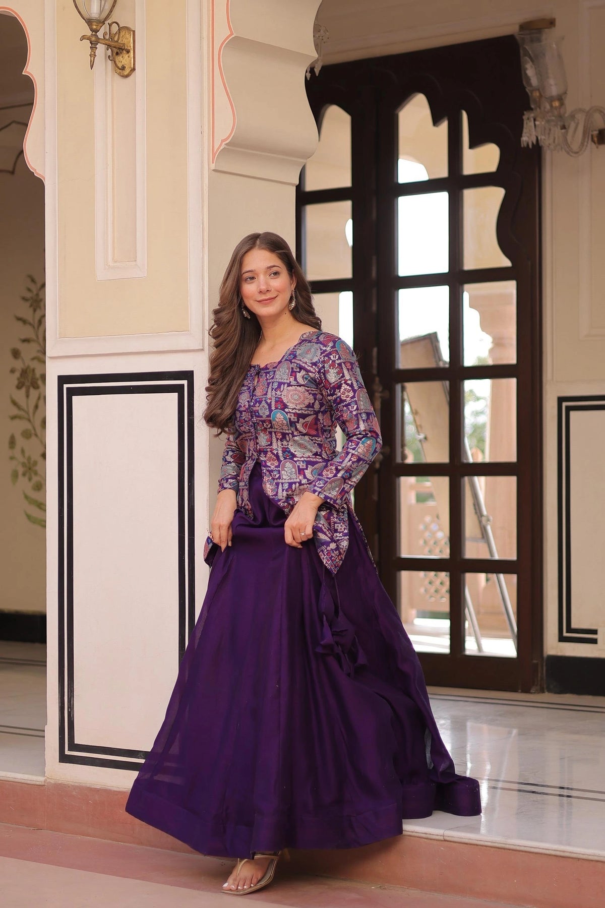 Woman in a purple floral top and long skirt standing in an ornate interior setting.