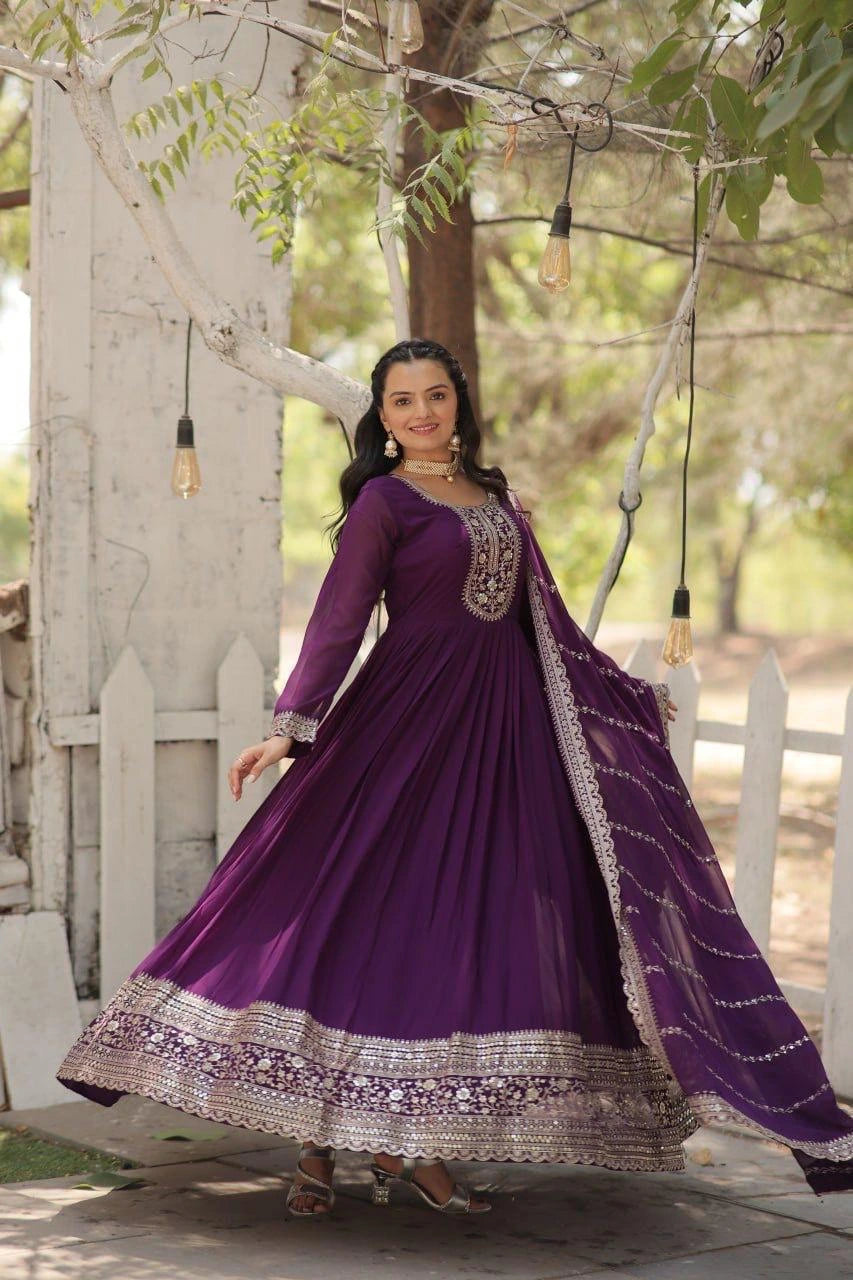 Woman in a purple traditional outfit standing outdoors with trees and a white fence in the background.
