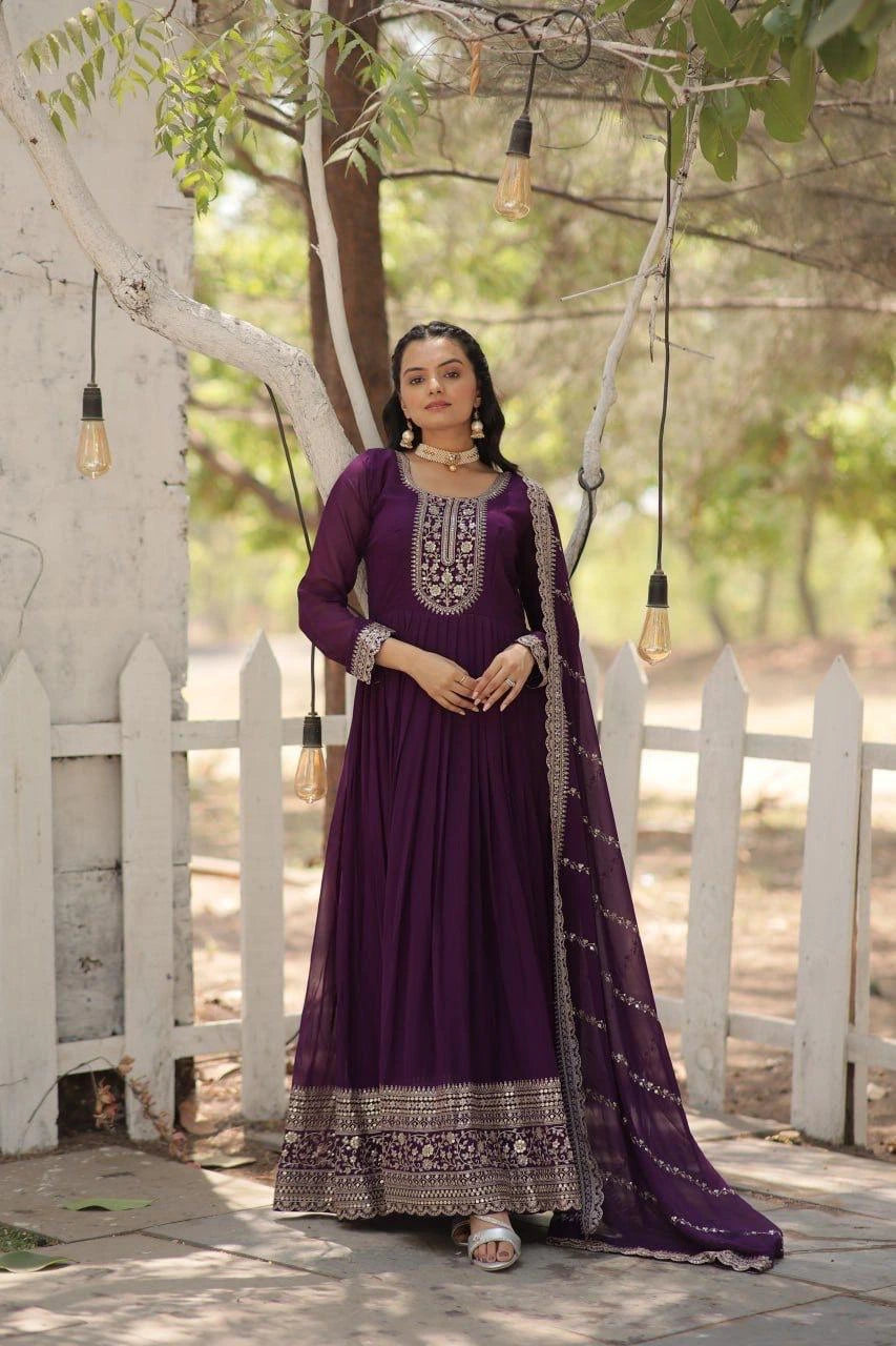 Woman in a purple traditional outfit standing outdoors with a white picket fence and greenery in the background.