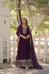 Woman in a purple traditional outfit standing outdoors with a white picket fence and greenery in the background.