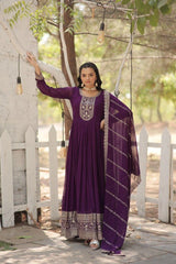 Woman in a purple traditional outfit standing outdoors with greenery and a white picket fence in the background.