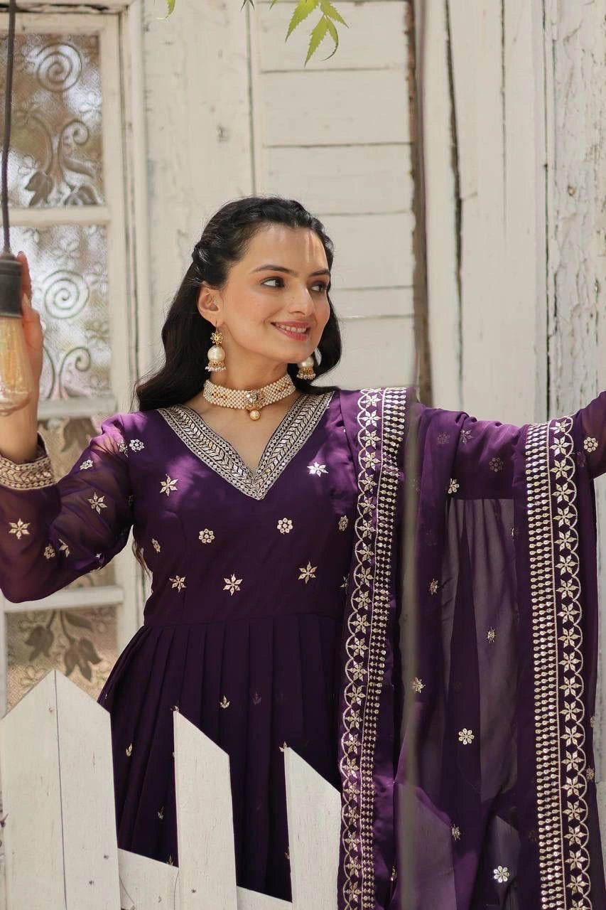 A woman wearing a deep purple traditional outfit with silver embroidery and a matching sheer dupatta, posing in front of a rustic white wooden door.