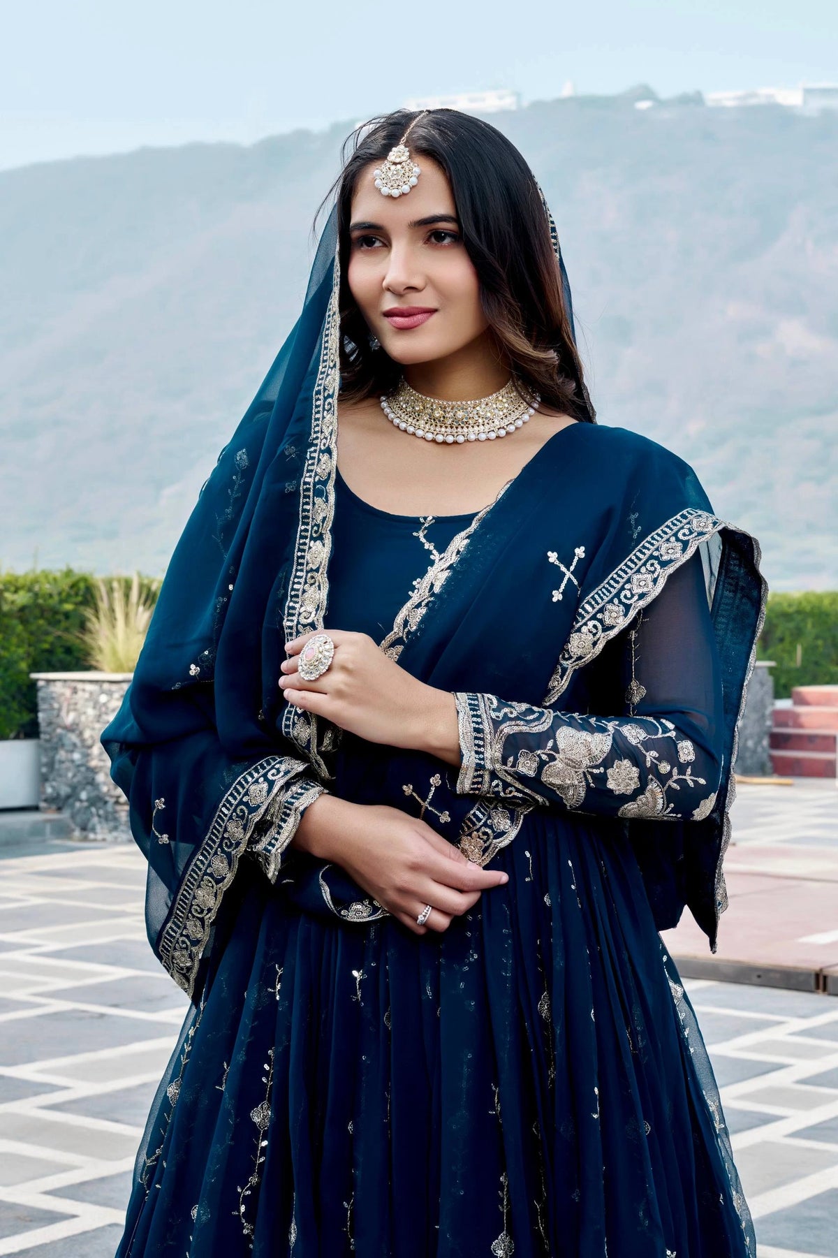 Woman in traditional dark blue outfit with embroidery, standing outdoors with mountains in the background.