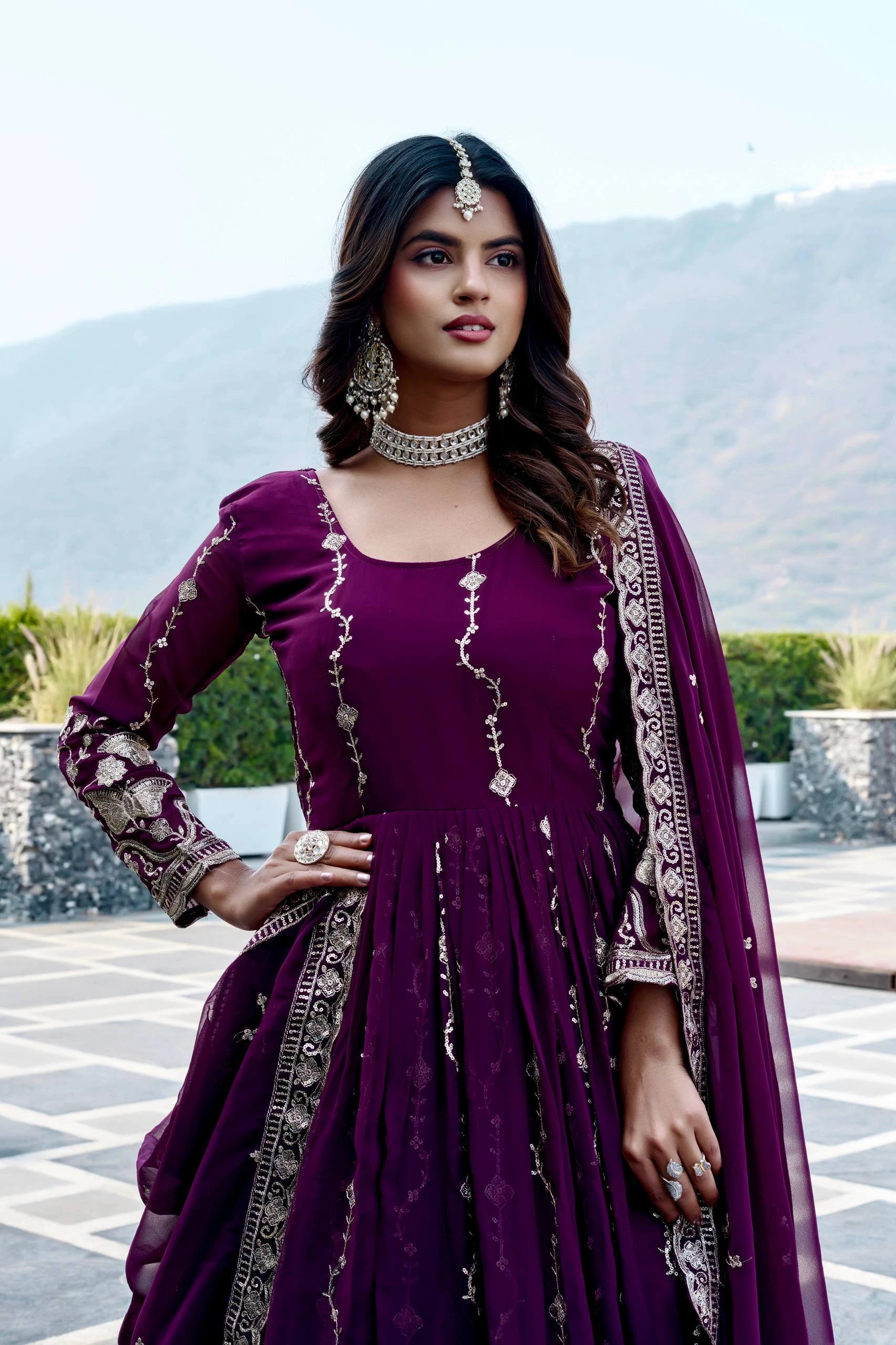 Woman in a purple traditional outfit with intricate designs, standing outdoors with mountains in the background.