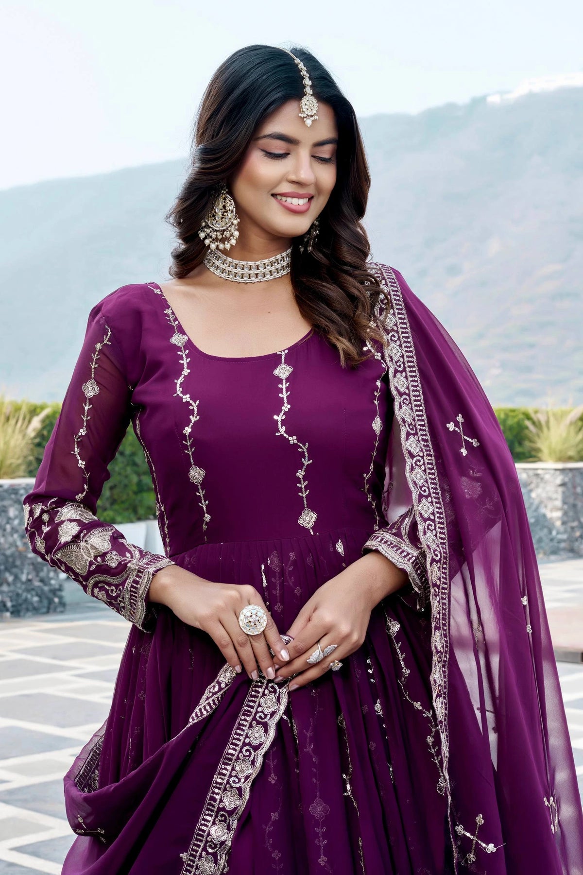 Woman in a purple traditional outfit with intricate designs, standing outdoors with mountains in the background.