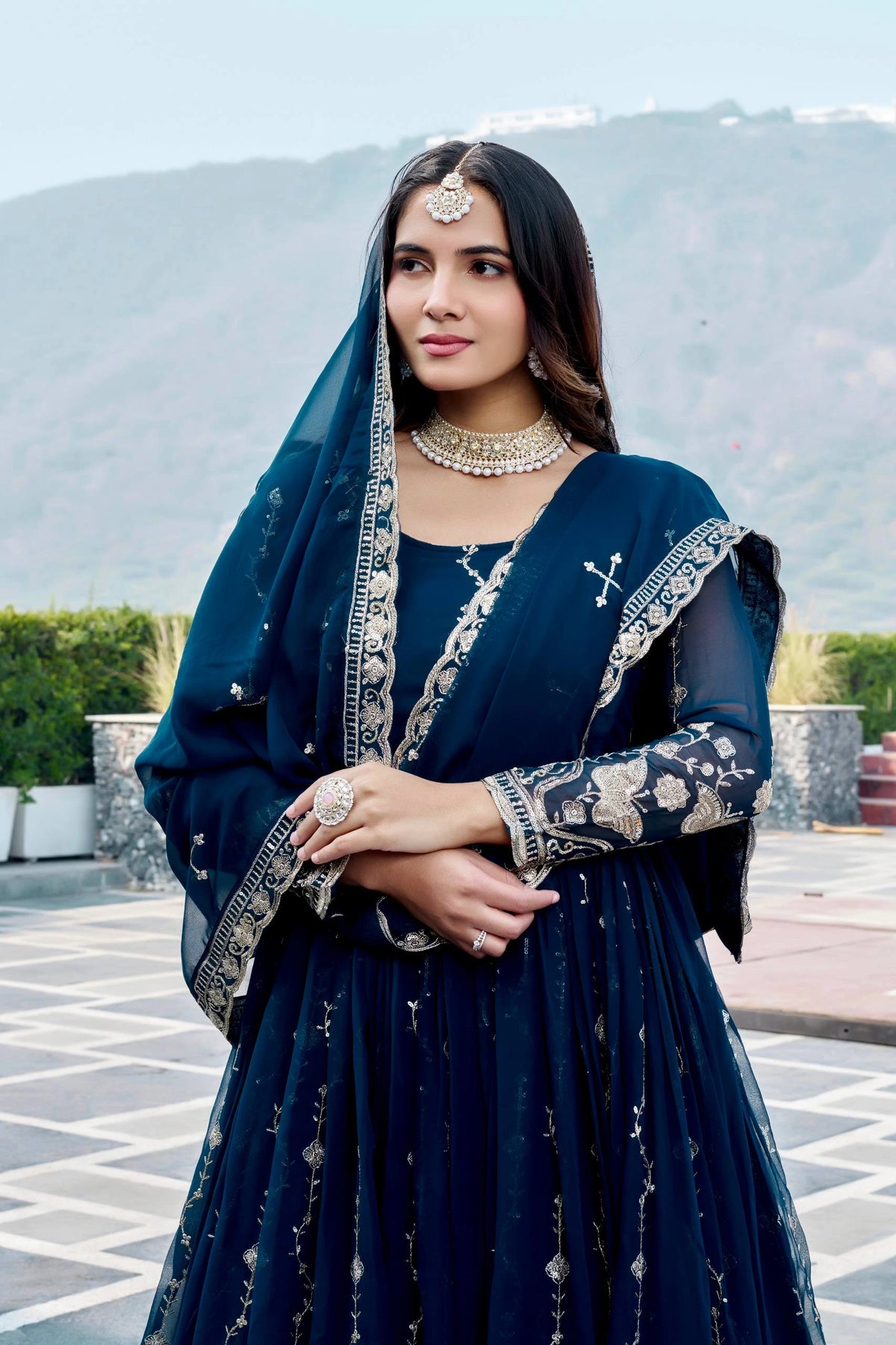 Woman in a dark blue traditional outfit with embroidery, standing outdoors with mountains in the background.