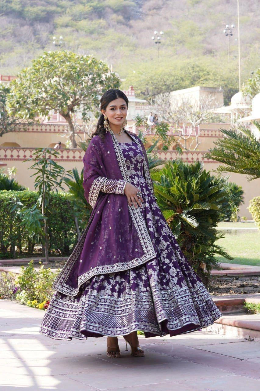 Woman in a purple traditional outfit standing outdoors with greenery in the background
