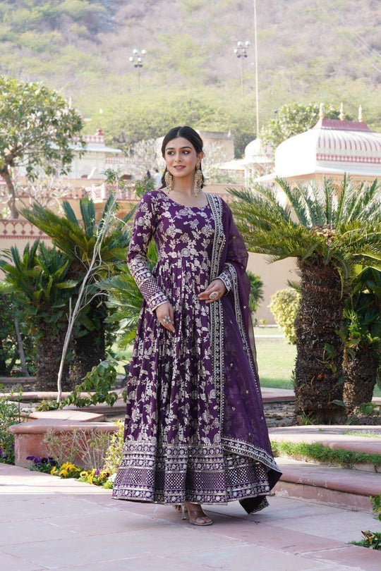 Woman in a purple traditional outfit standing in a garden with greenery and architectural elements in the background.