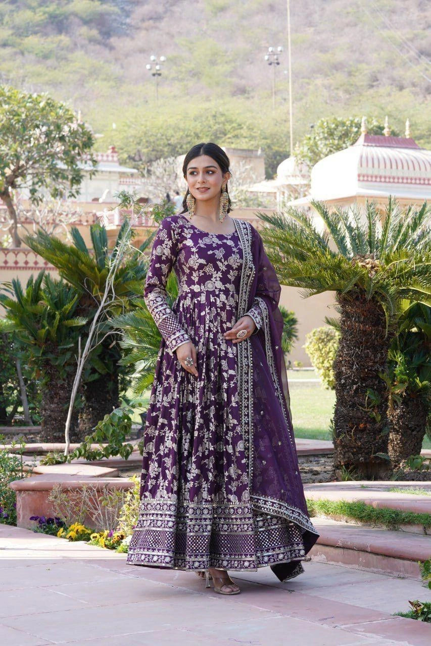 Woman in a purple traditional outfit standing in a garden with greenery and architectural elements in the background.
