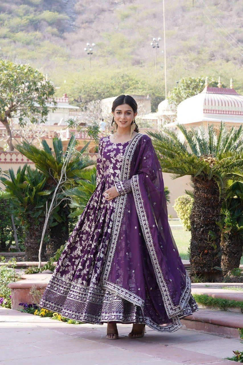 Woman in a purple traditional outfit standing outdoors with greenery and architecture in the background