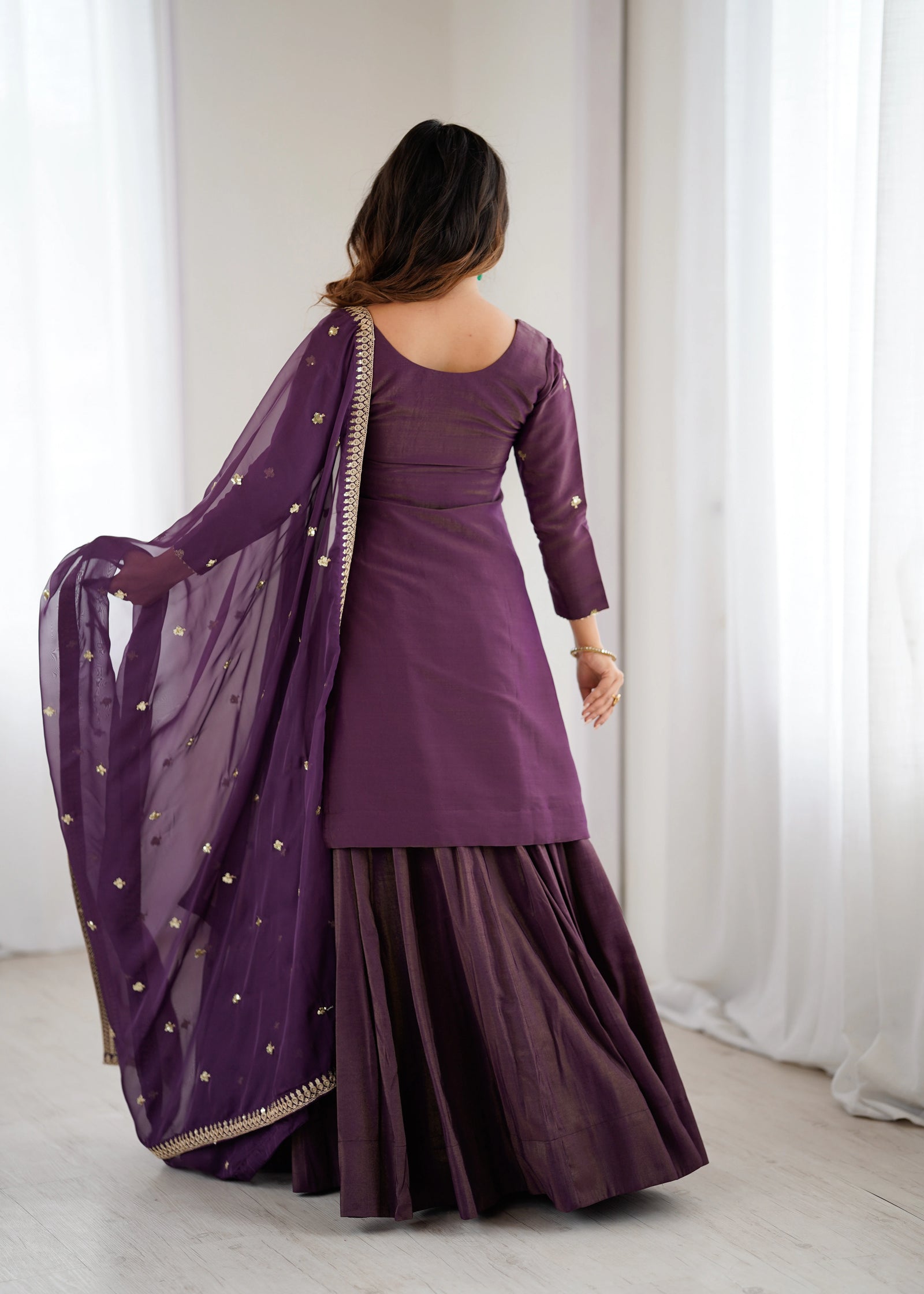 Woman wearing a purple traditional outfit with a sheer dupatta against a white curtain background