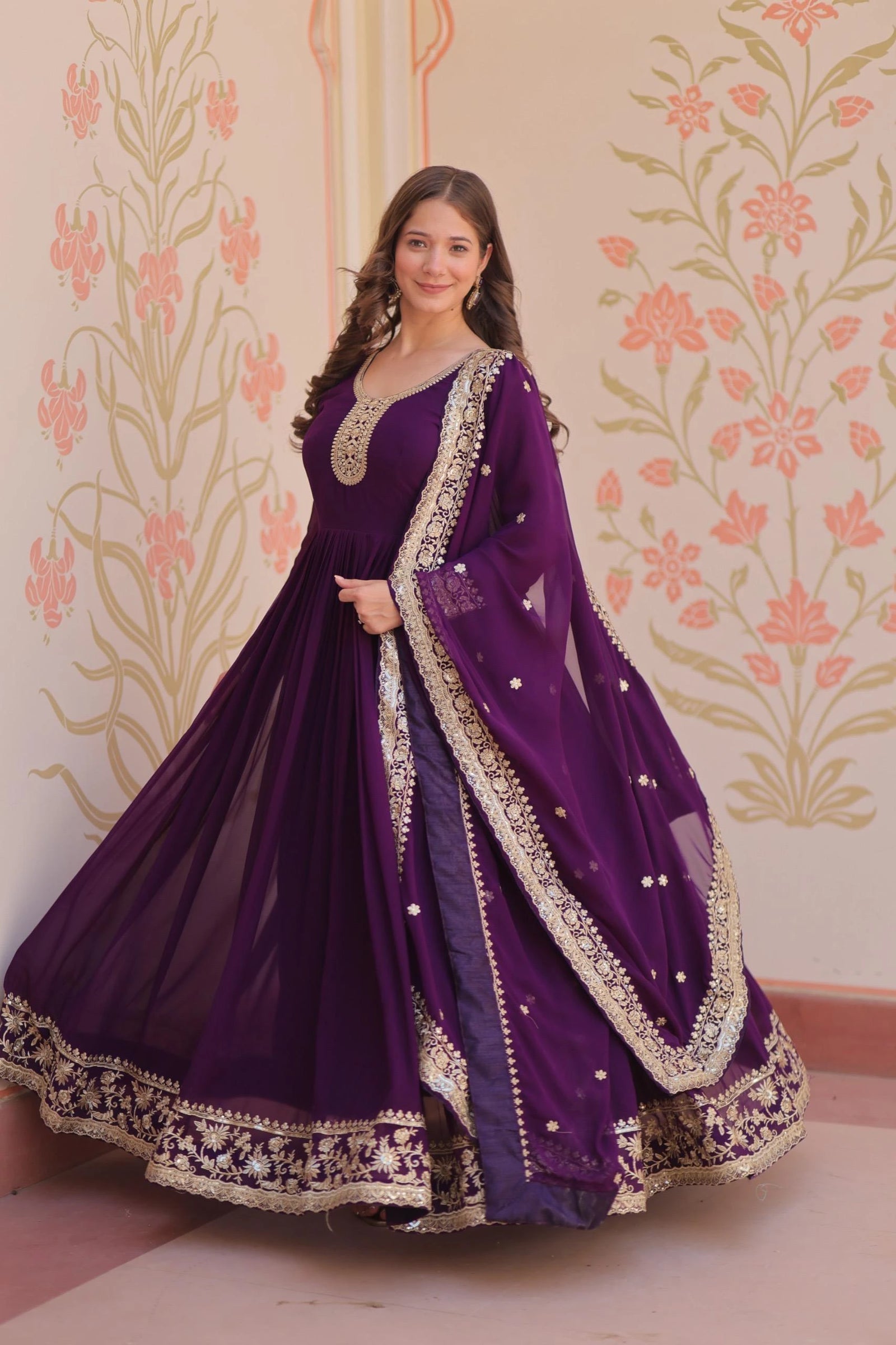 Woman in a purple traditional outfit with gold embroidery against a floral wallpapered wall.