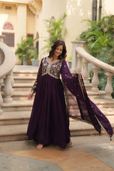Woman in a purple and gold traditional outfit standing in an elegant indoor setting with columns and plants.