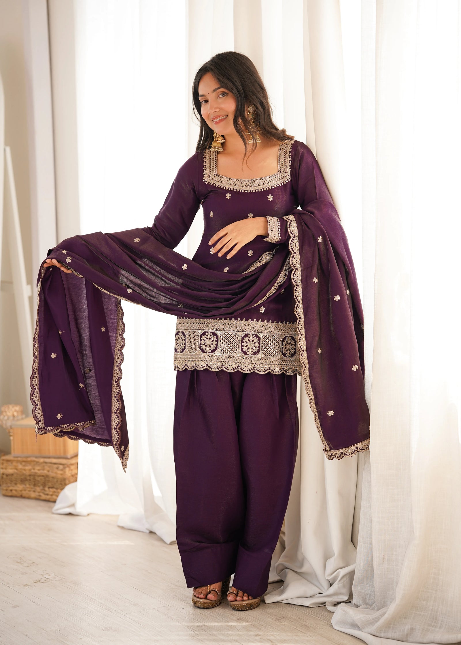 Woman in a traditional outfit with a purple dress and dupatta against a white curtain background