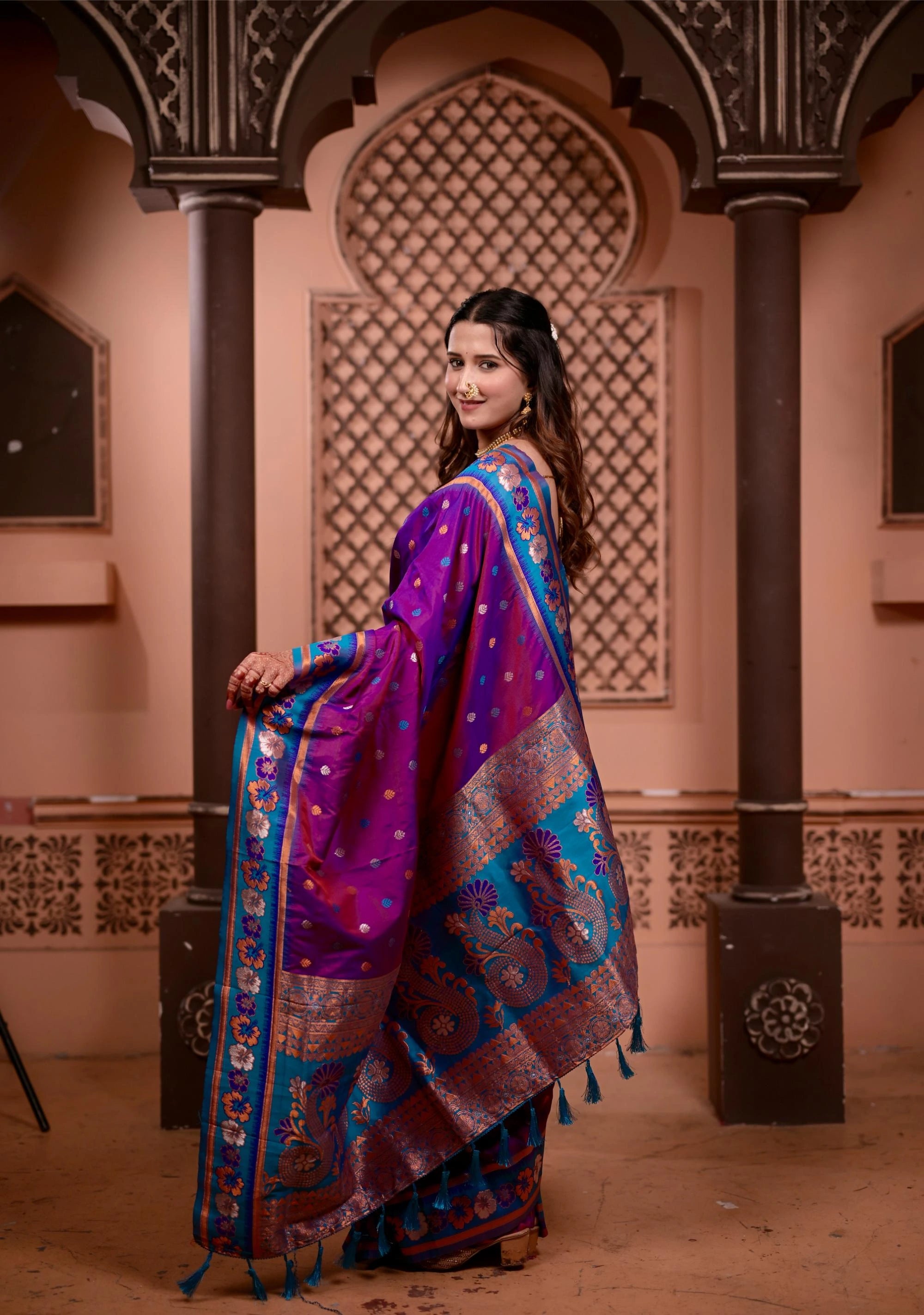 Woman in a traditional purple and blue saree standing in an ornate architectural setting.