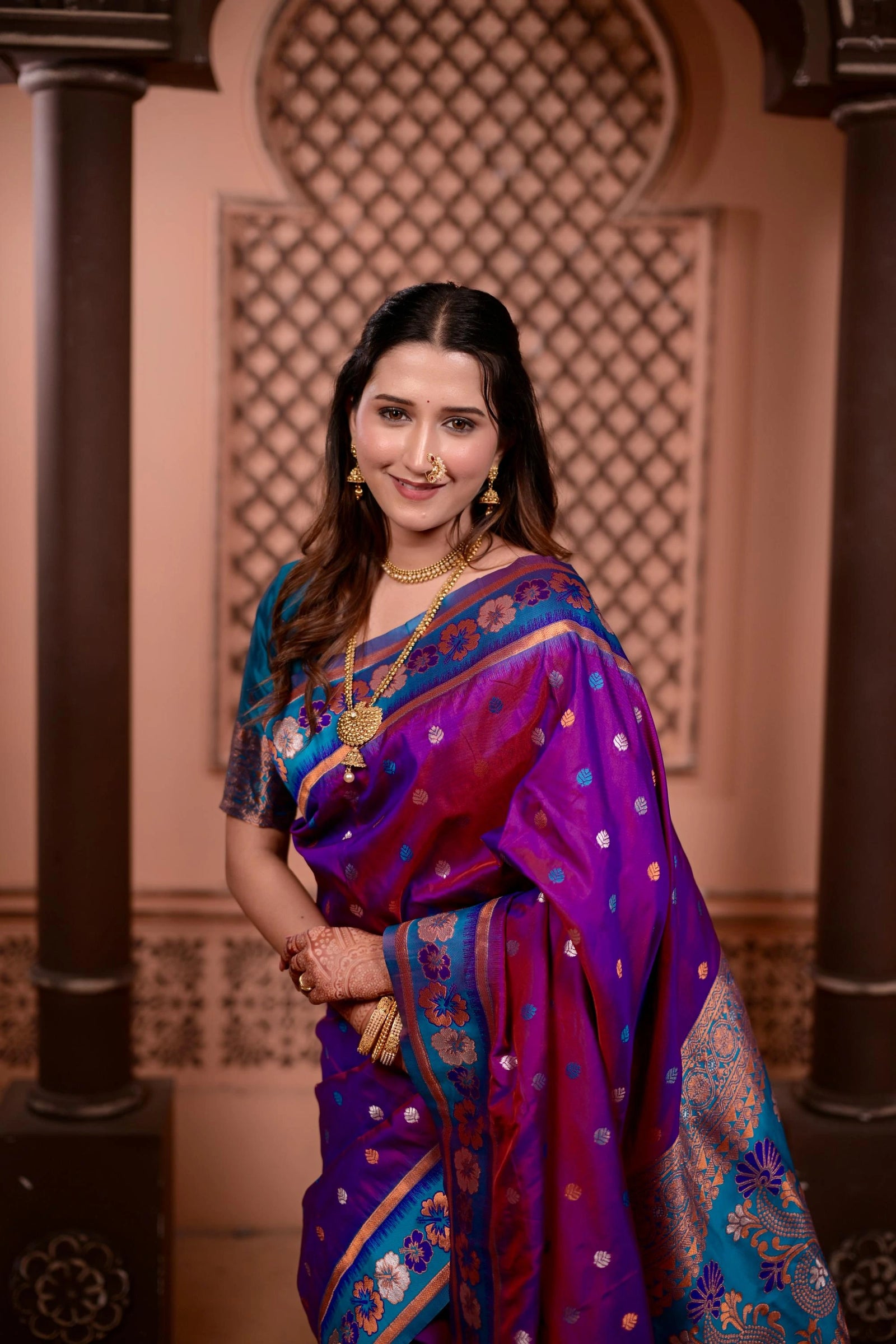 Woman in a traditional purple and blue saree standing in an ornate indoor setting.