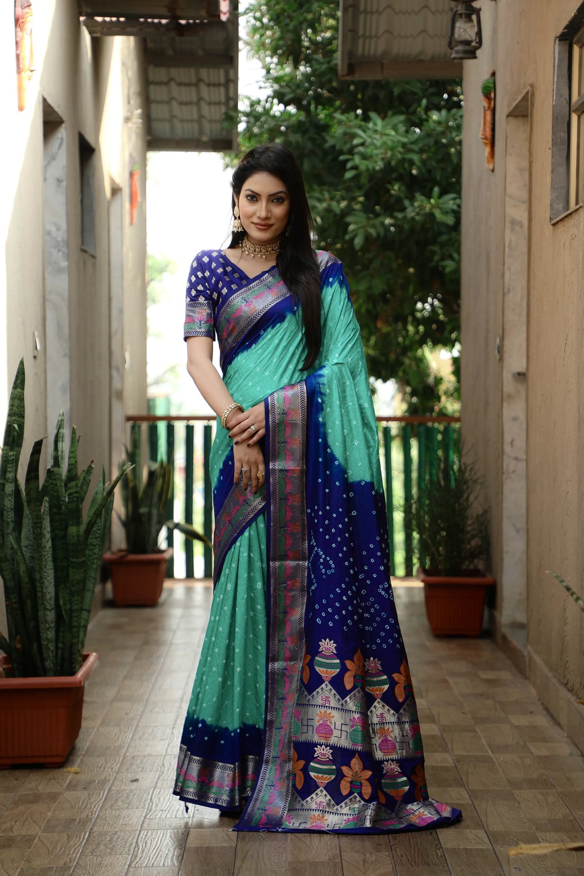 Woman wearing a colorful saree in an outdoor setting with plants and a building.