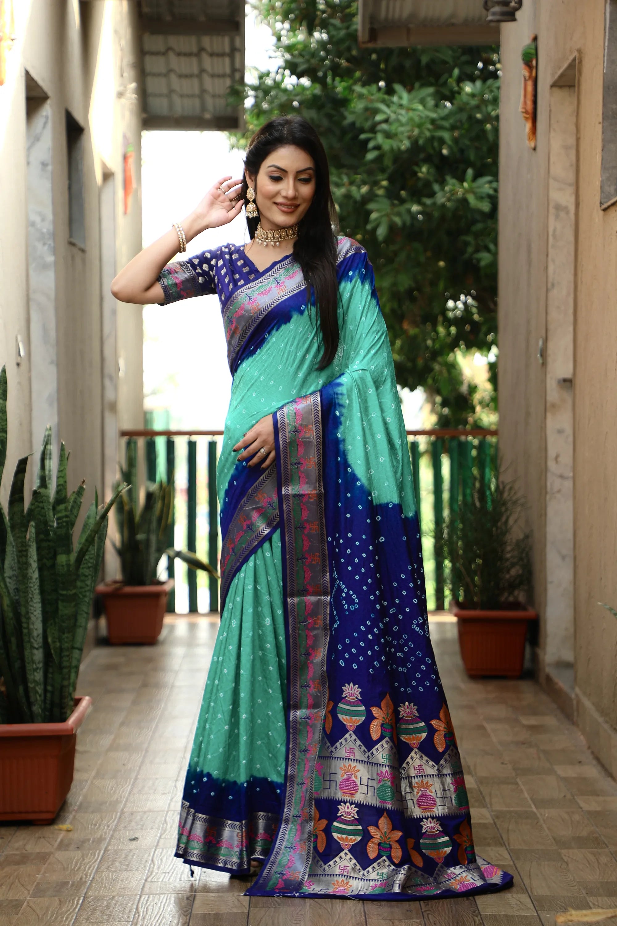 Woman wearing a colorful saree in an outdoor setting with plants and a building.