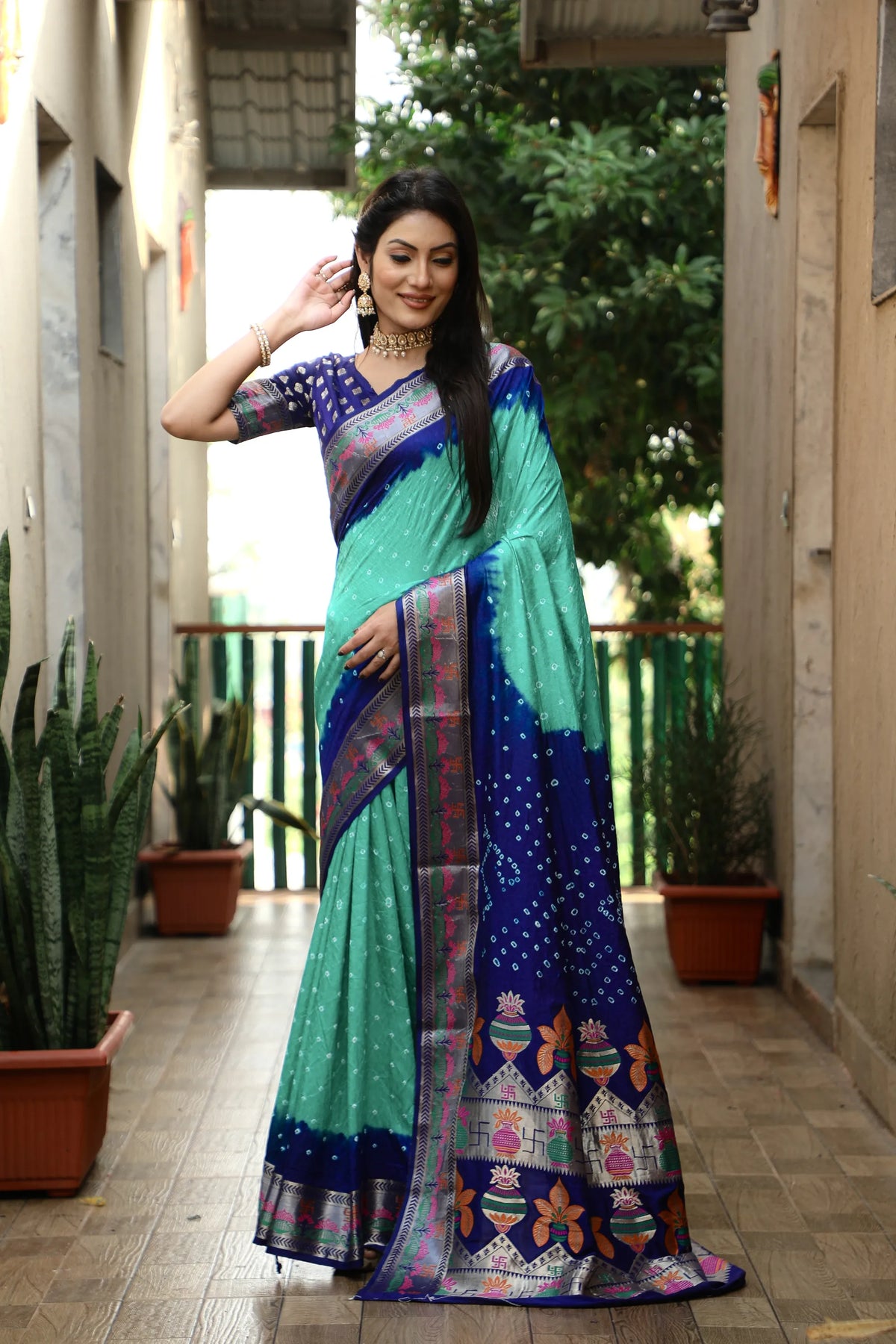 Woman wearing a colorful saree in an outdoor setting with plants and a building.