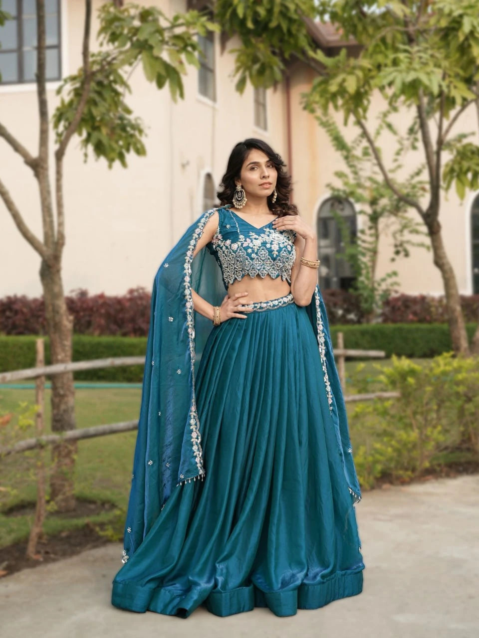 Woman in a blue traditional outfit standing outdoors with greenery in the background