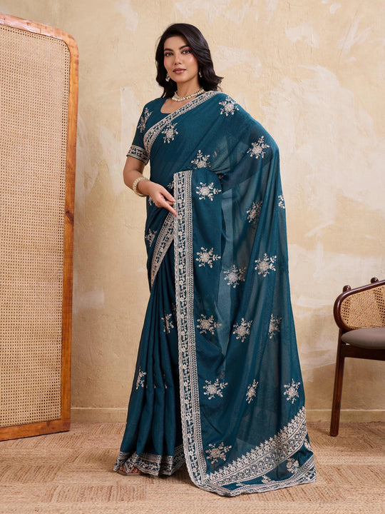 Woman wearing a teal saree with silver embroidery against a beige wall.