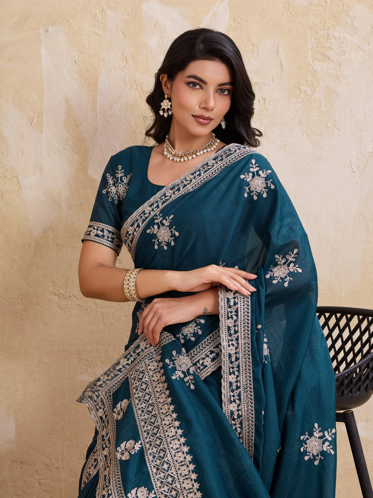 Woman wearing a teal saree with silver embroidery against a beige wall.