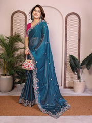 Woman wearing a blue saree with a floral design, standing indoors with plants in the background.