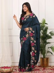Woman wearing a dark blue saree with floral patterns in a room with white walls and a plant.