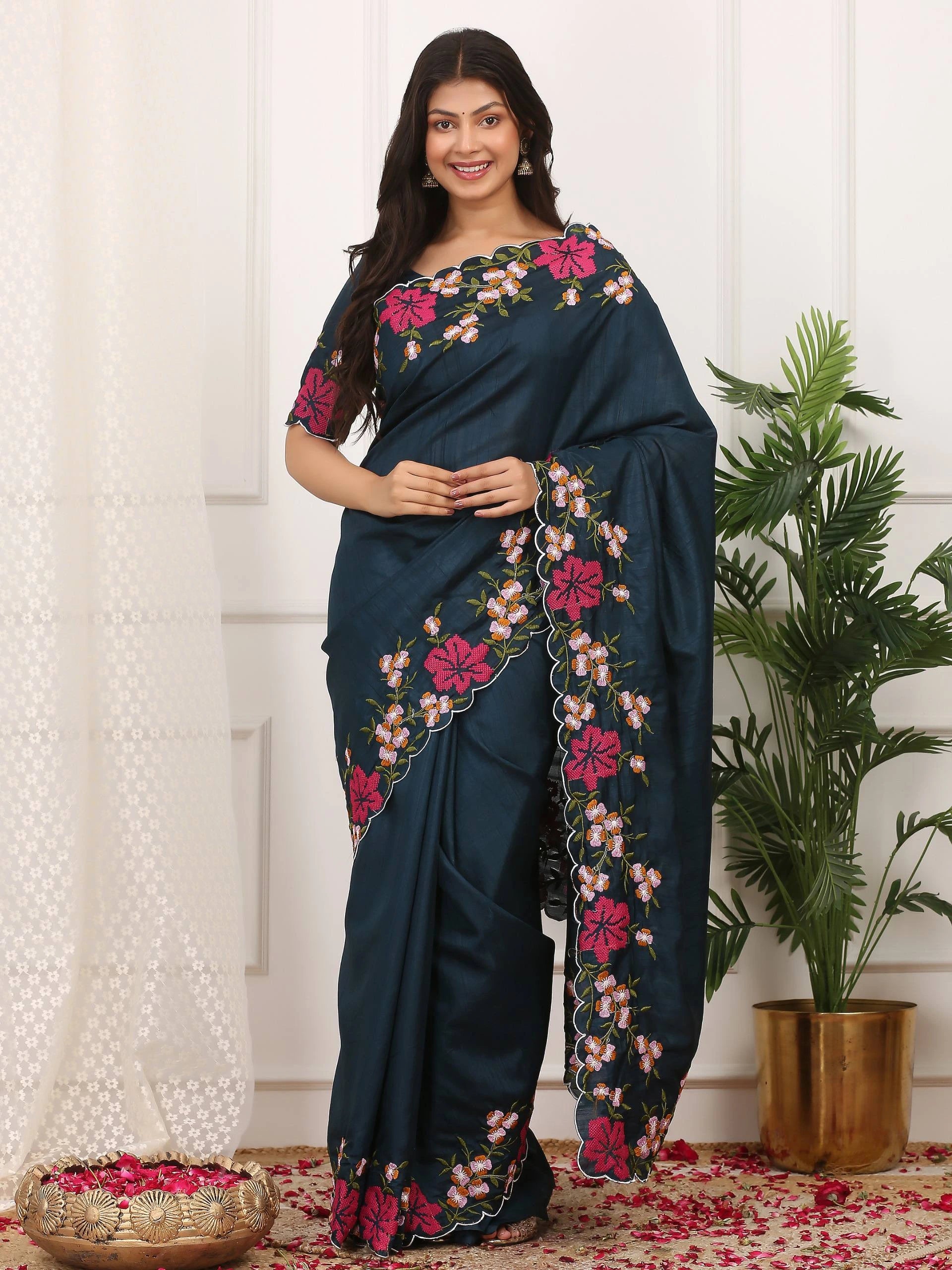 Woman wearing a dark blue saree with floral embroidery in a room with a plant and decorative items.