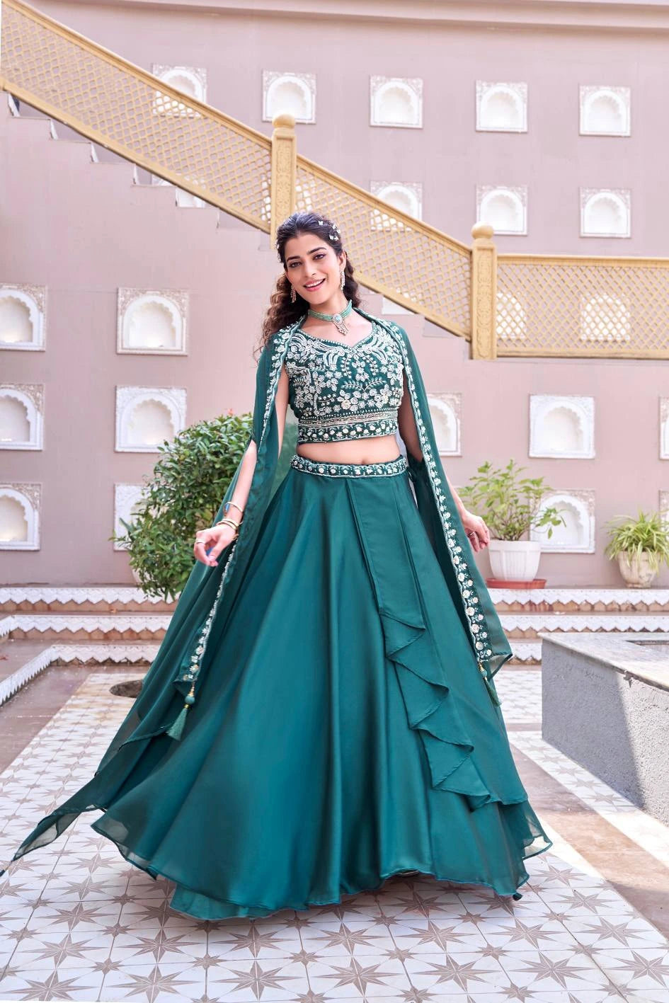 Woman in a green traditional outfit standing in an outdoor setting with plants and architectural elements.