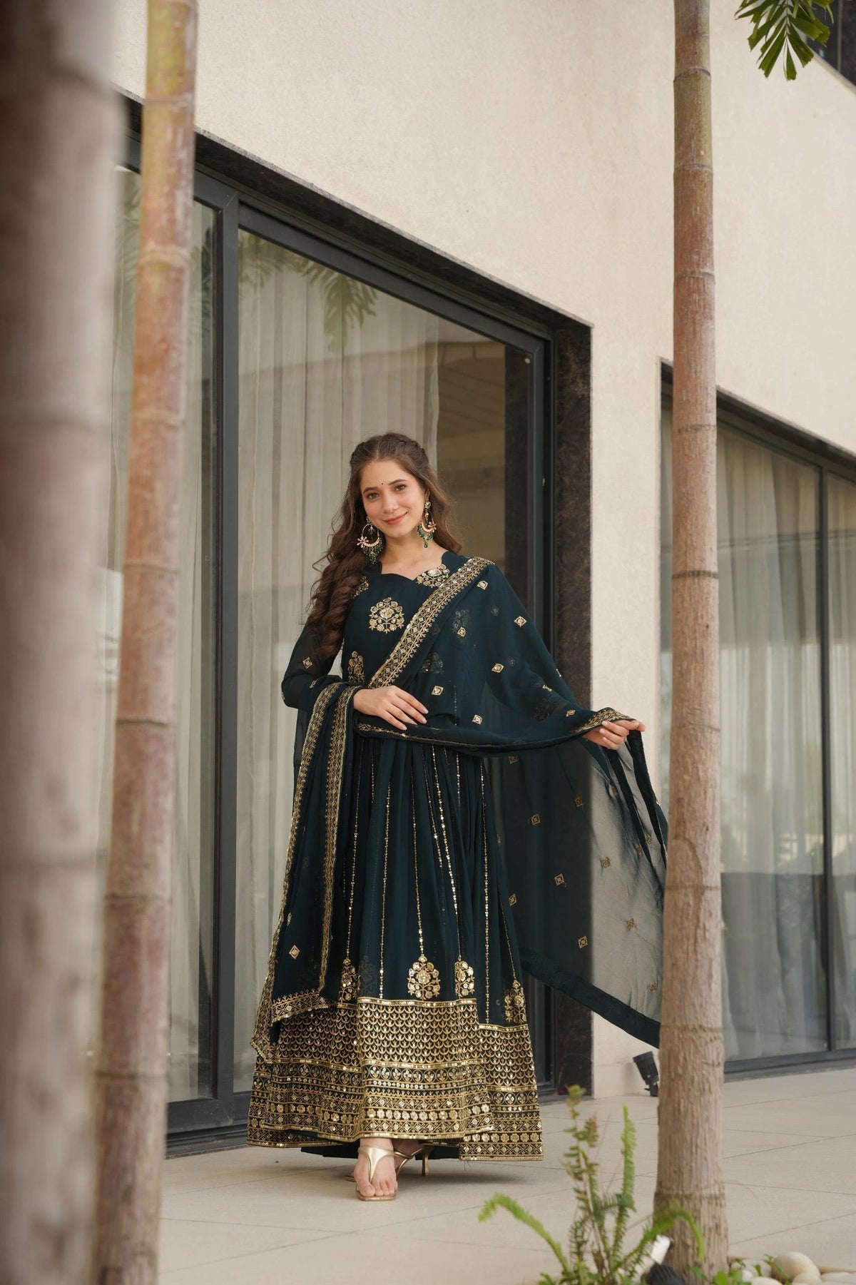 Woman in a dark green traditional outfit with gold details standing in front of a modern building.