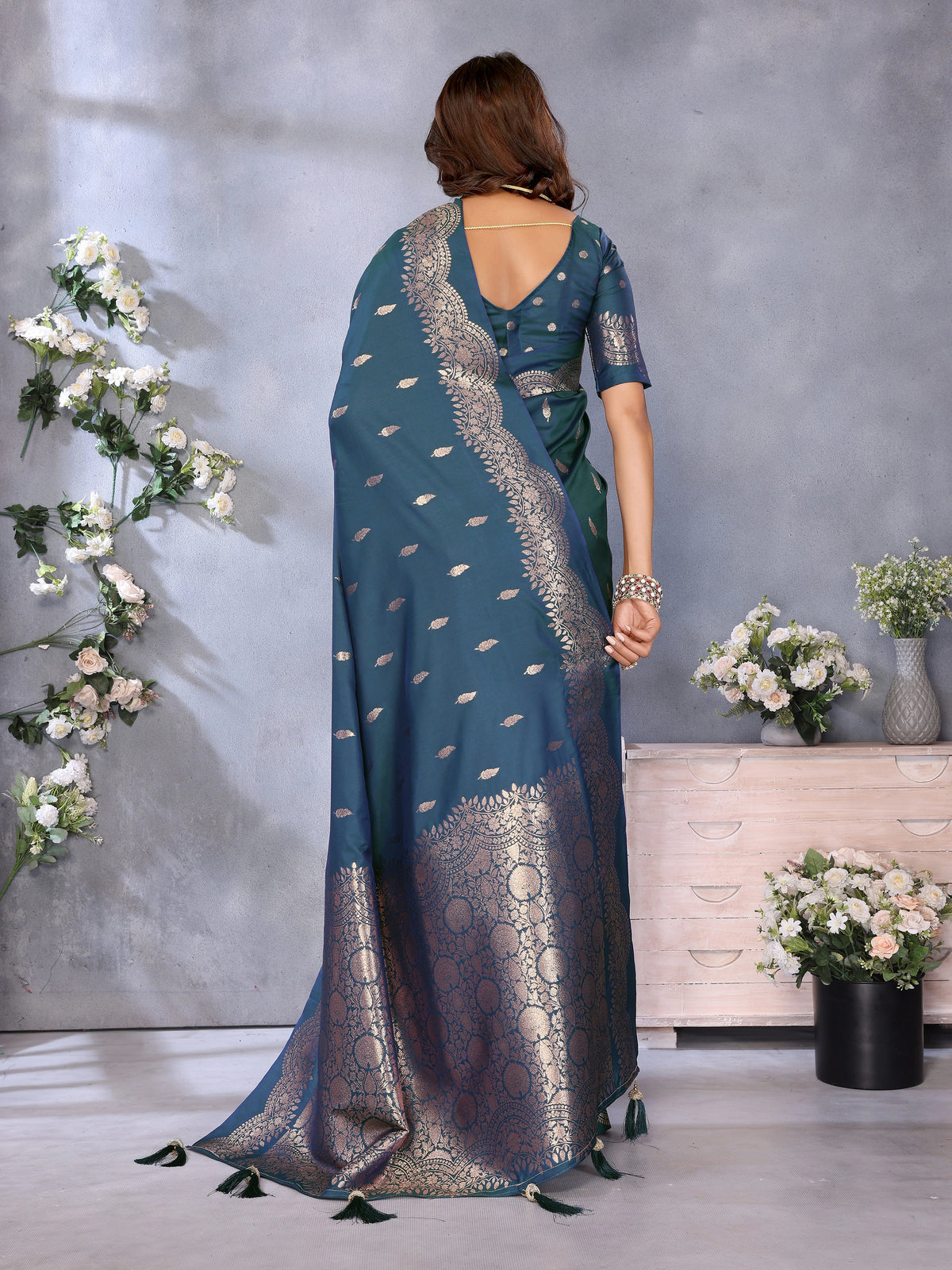 Woman wearing a blue saree with silver patterns in a room with decorative flowers and furniture.
