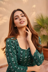 A medium shot of a woman in a dark green dress with white patterns, smiling and resting a finger on her chin in a sunlit outdoor setting.