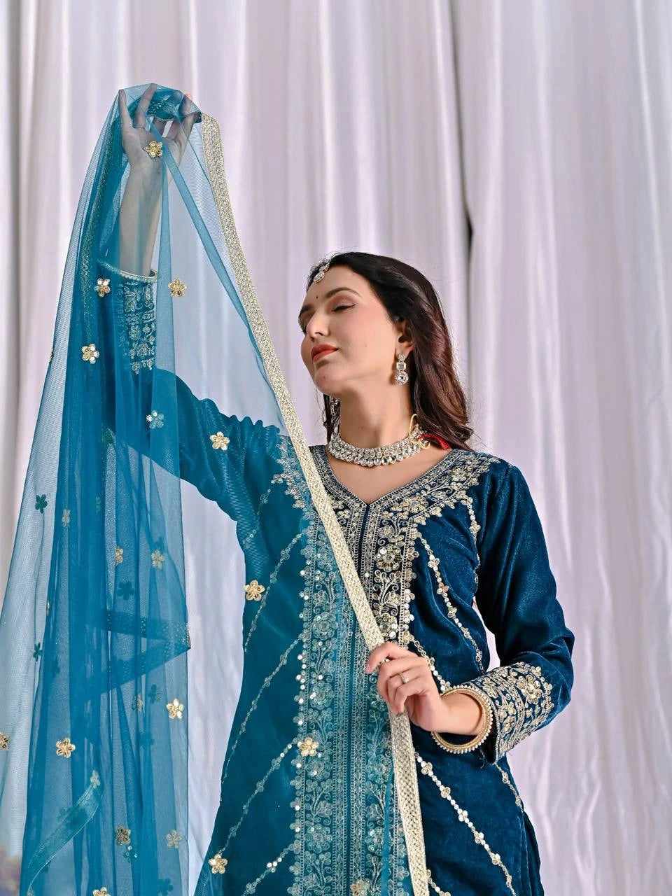 Woman wearing a blue embroidered traditional outfit with a matching dupatta against a white curtain background