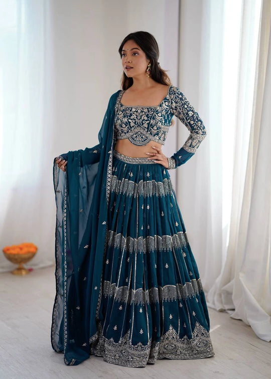 Woman in a traditional teal and silver outfit standing in a bright room with white curtains.