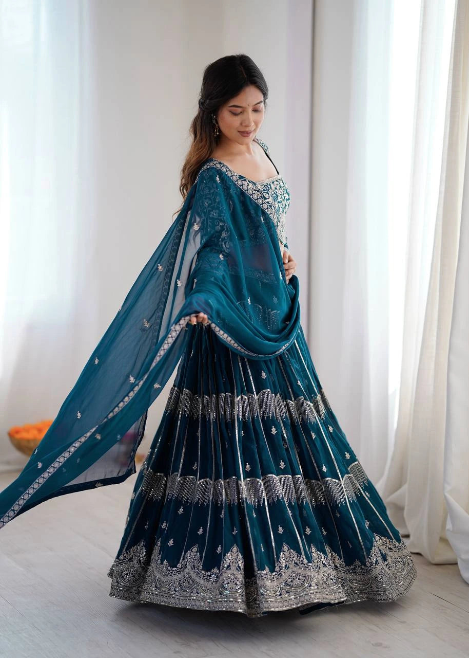 Woman in a teal and silver traditional outfit standing against a white curtain.