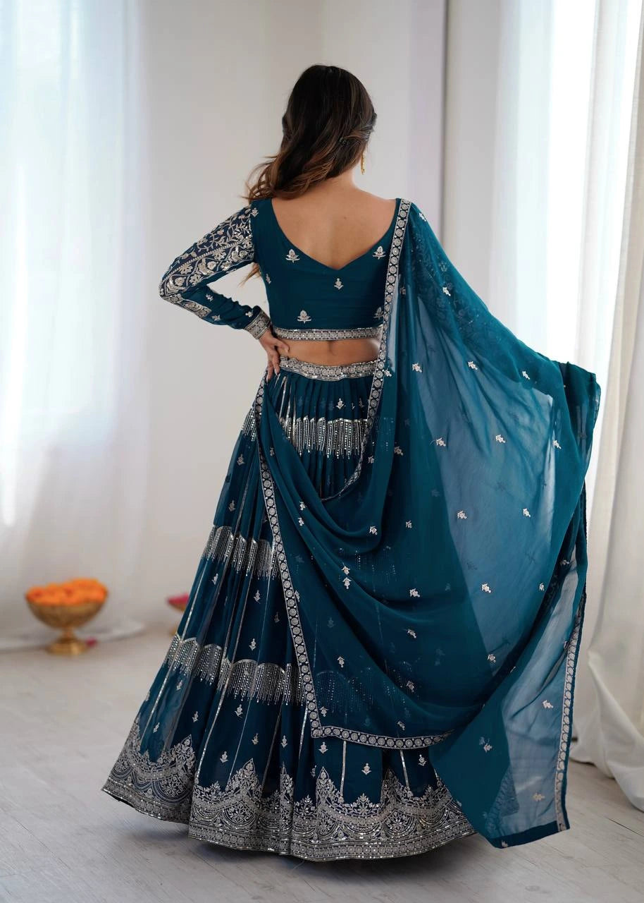 Teal embroidered lehenga with silver details worn by a person, standing against a white curtain background.