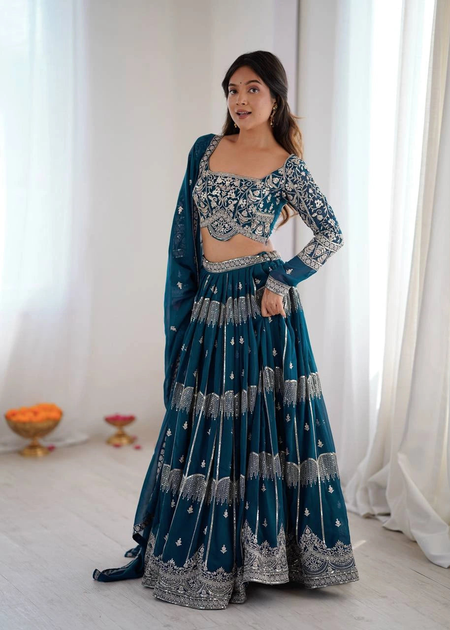 Woman in a blue and silver traditional outfit standing in a bright room with white curtains.