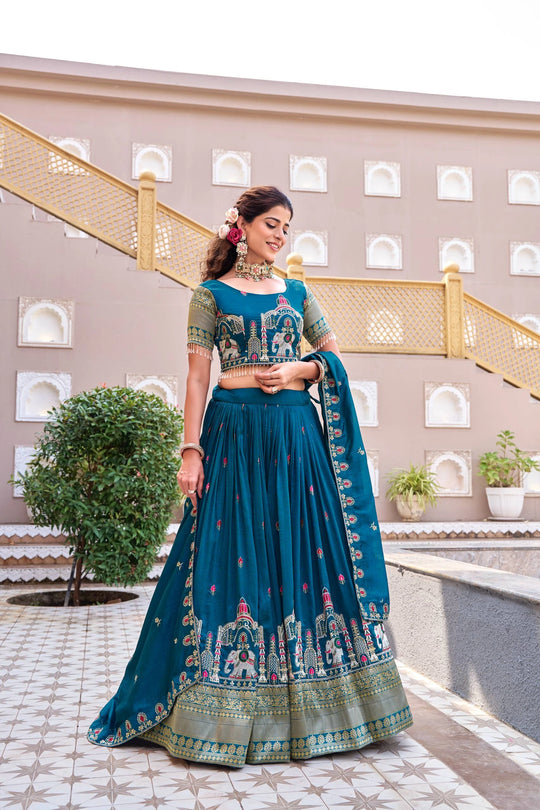 Woman in a traditional teal and green outfit standing in front of a decorative building.