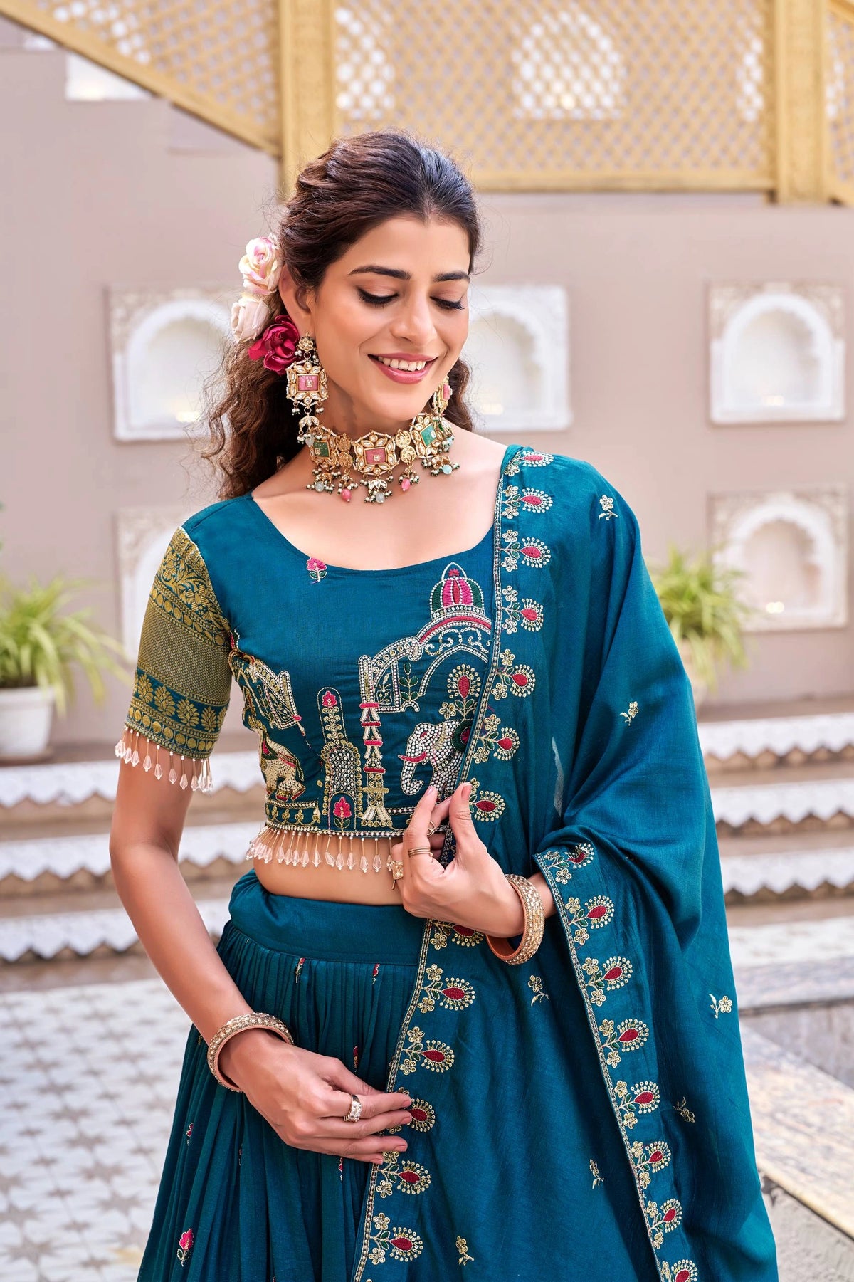 Woman wearing a traditional blue embroidered outfit with jewelry, standing against an architectural background.