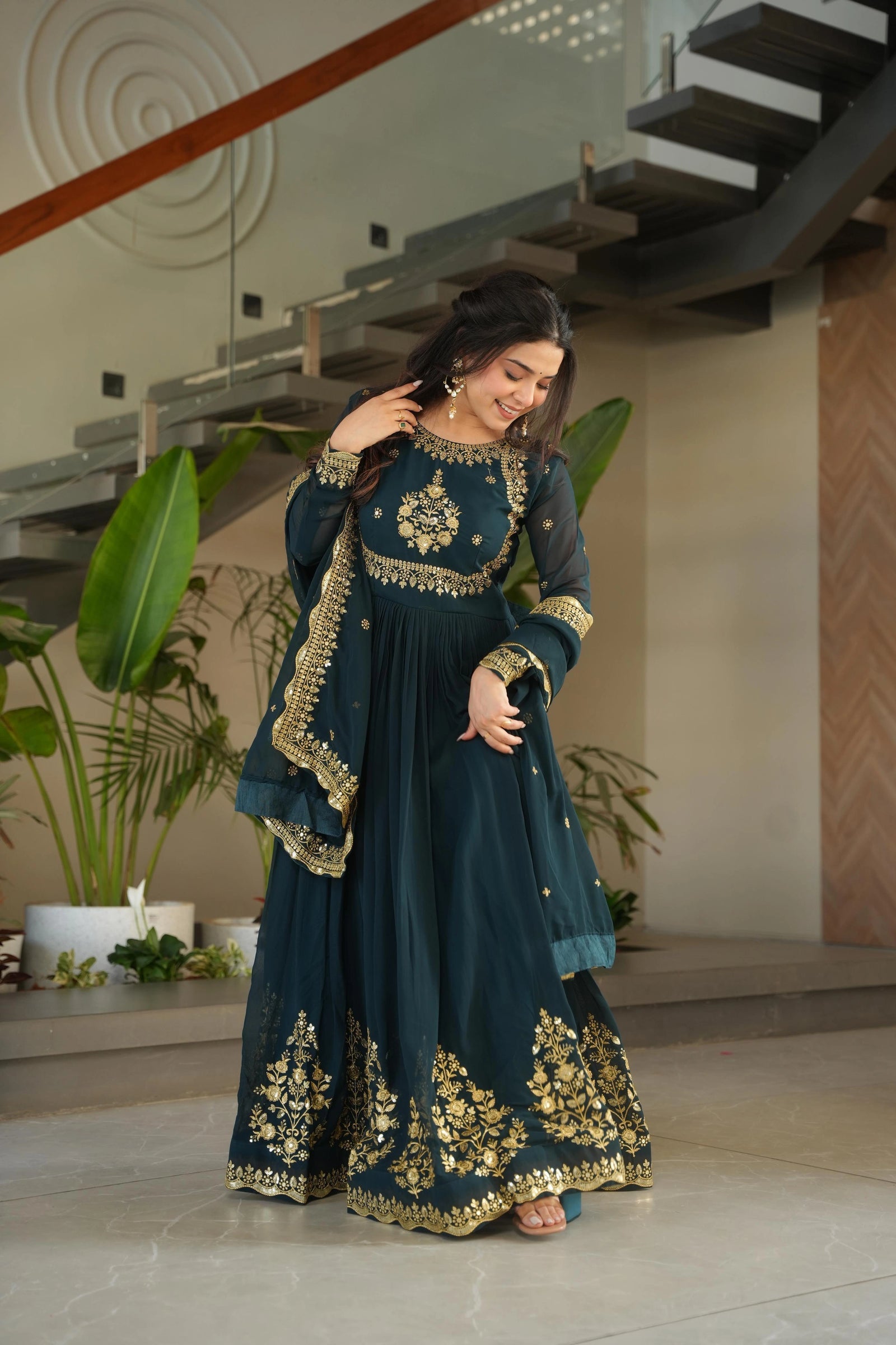 Woman in a teal and gold traditional outfit standing indoors with plants and a staircase in the background.