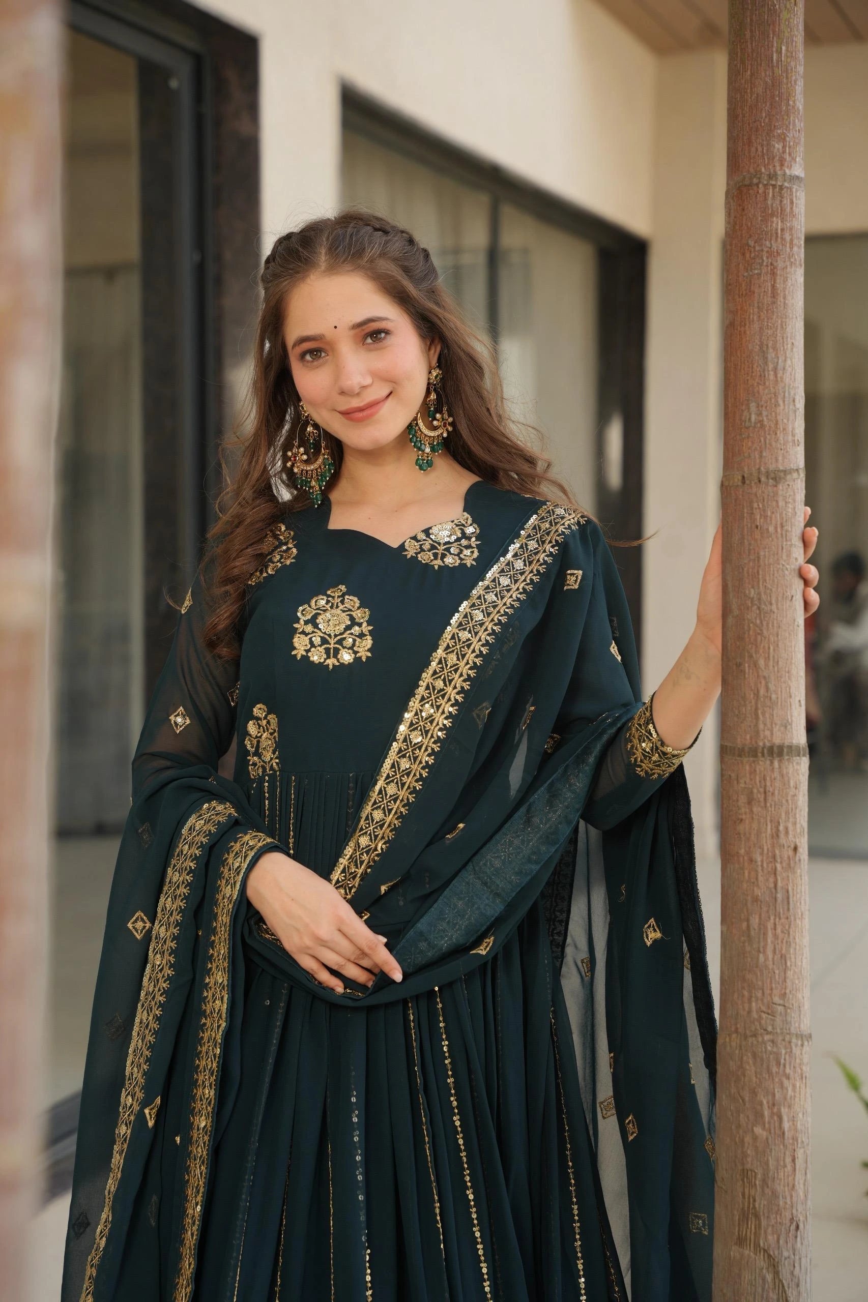 Woman in a dark green traditional outfit with gold embroidery standing against a building.