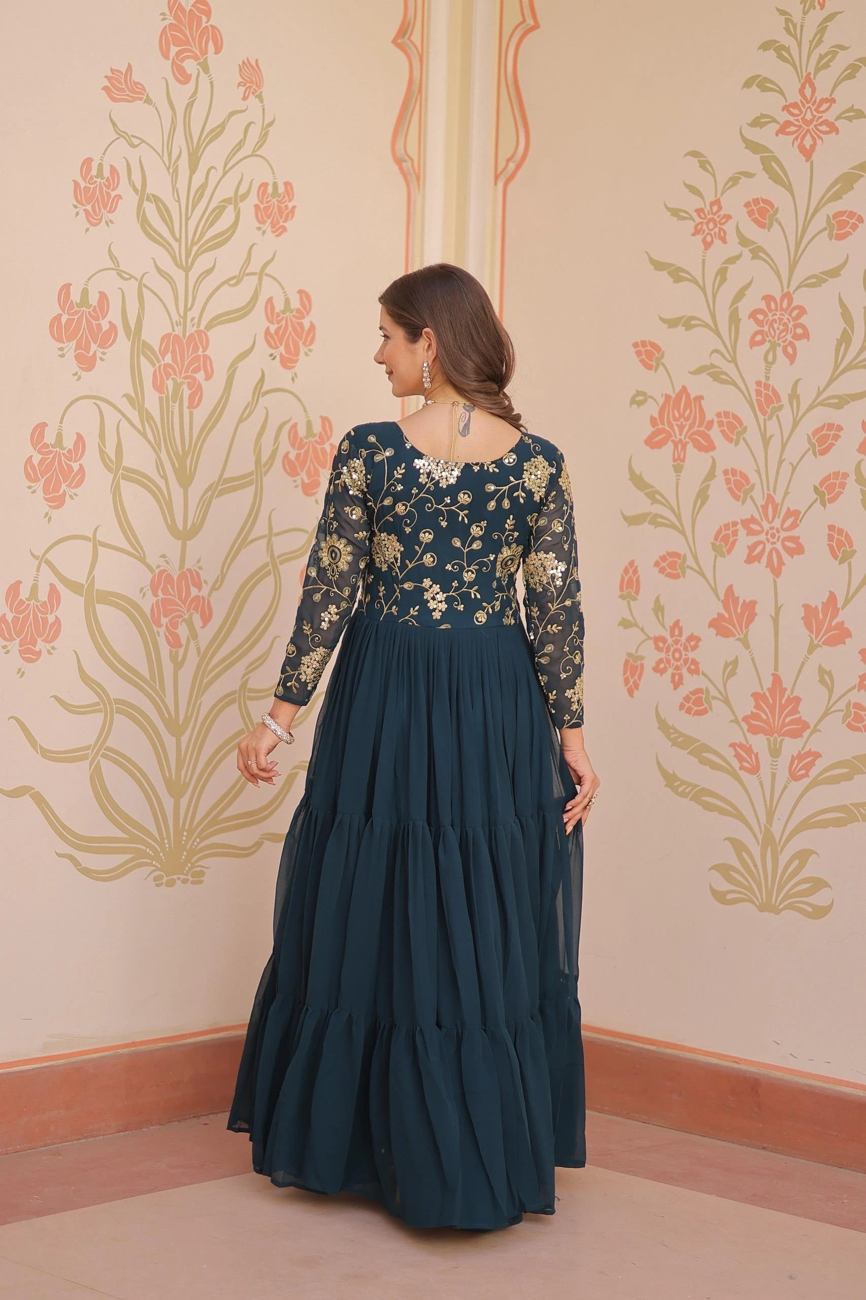 Woman in a dark blue traditional outfit with floral patterns against a decorative wall.