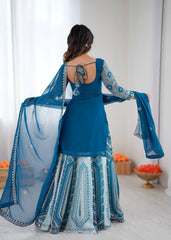 Woman wearing a blue traditional outfit with a white and blue patterned dupatta, standing against a white wall.