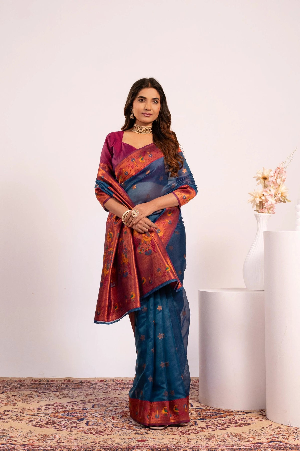 Woman wearing a colorful saree standing against a white wall with a vase of flowers.